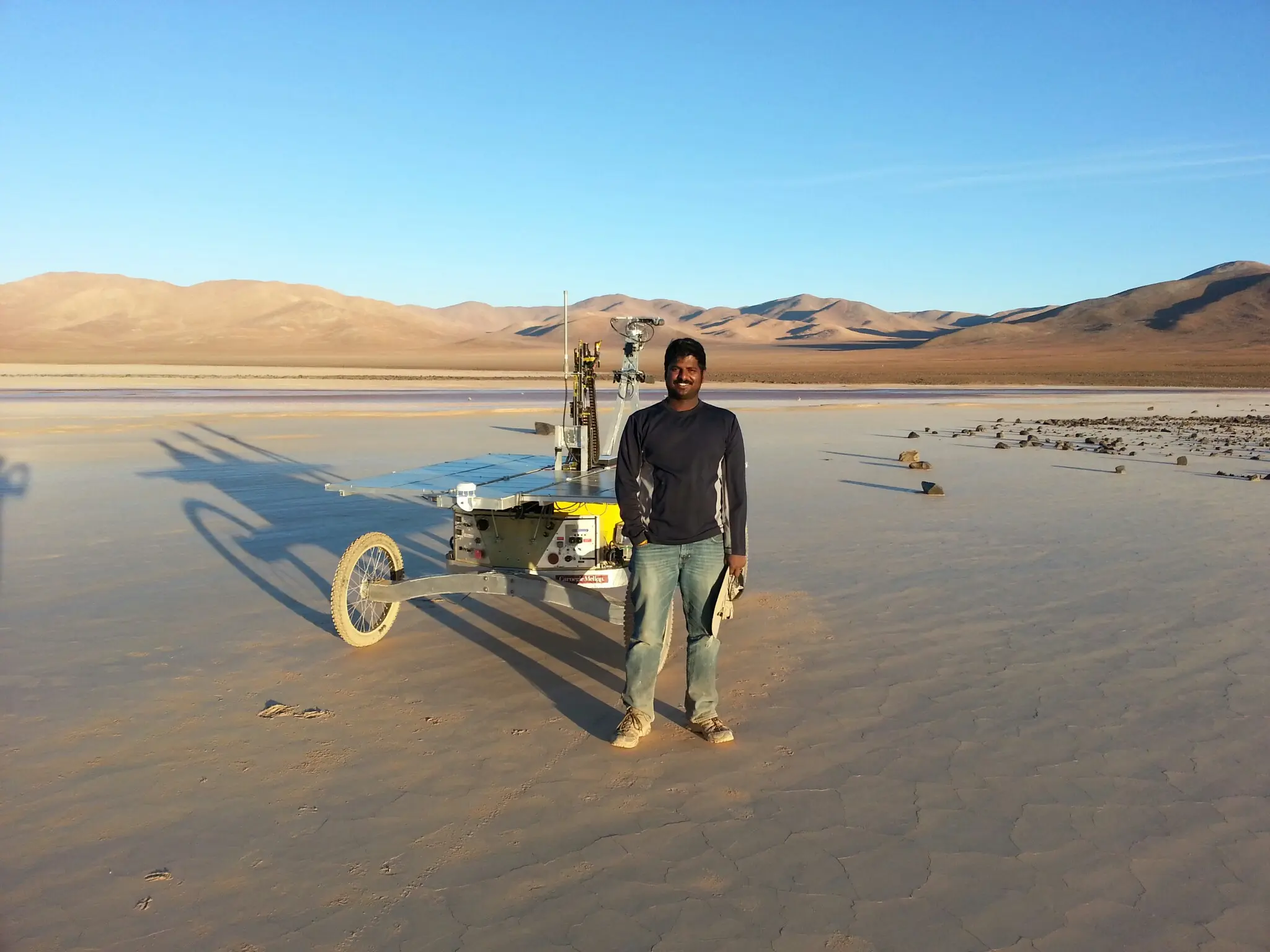 Srini Vijay standing beside a Carnegie Mellon solar-powered field rover on a dry salt flat, mountains behind.